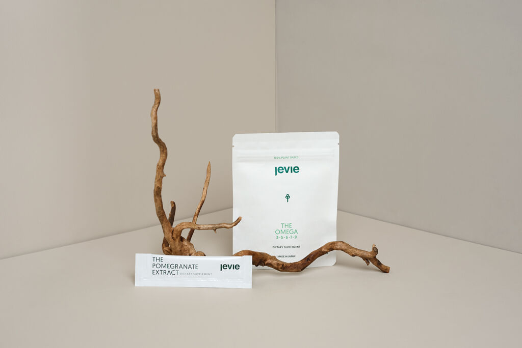 Two walls with a corner behind the product, Behind a branch the Omega package is standing. In front of the branch a pouch from the Pomegranate Extract is standing.