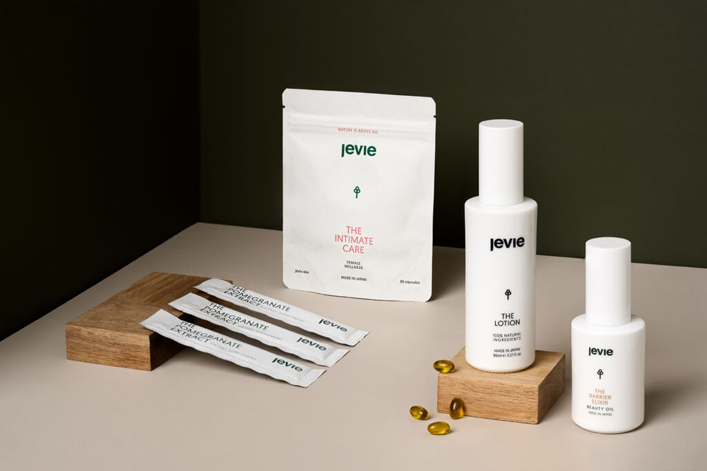 A cornered scene with both dark green walls, slightly from above with a beige background. All the products are shown. On the left is a wooden plateau where the pomegranate extract leans onto. Behind it is the intimate care and in the foreground there is a small wooden plateau where the lotion is standing on, but also some pills are leaning onto. Right from that plateau is the barrier elixir. 