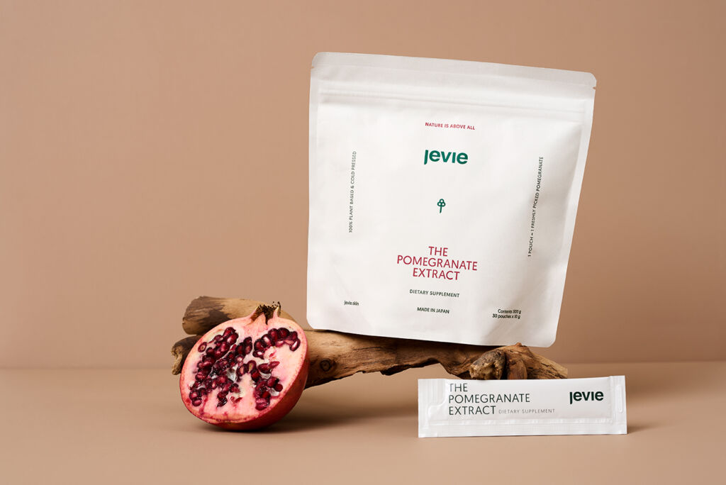 A copper background with a bark with the pomegranate extract bag on it. Next to it is a fresh cut in half pomegranate and in front is a small pouch from the package. 