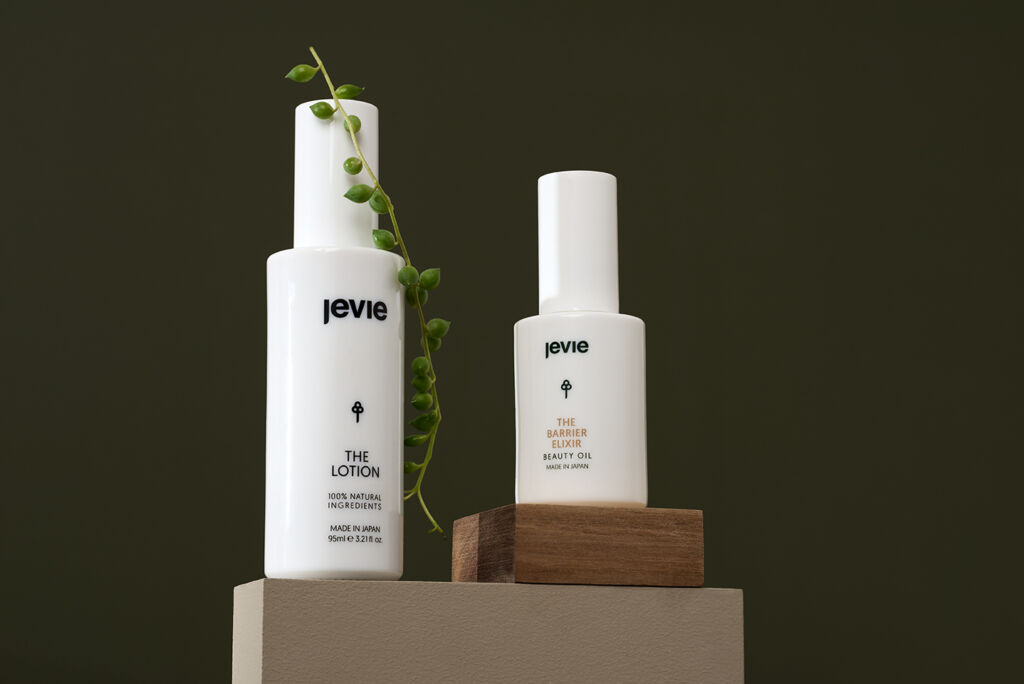 Green background with a rectangle block in a taupe colour. On the left is  a white glass bottle with a pea plant hanging on it. On the right is a smaller white glass bottle standing on top of a small wooden block.