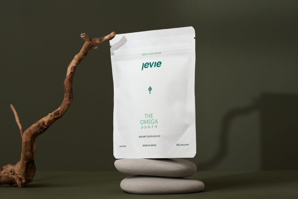 Green background, the packaging is white and standing on two grey rocks. Onto the packaging is a bark leaning, which creates a small shadow onto the product. 