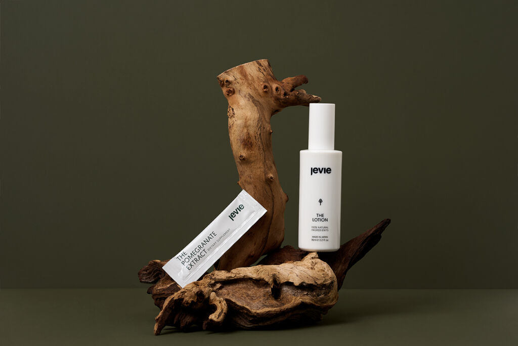 Green background with an wooden bark standing on it. On the right side on top of the bark is The Lotion, which is a white package. On the left-hand side, there is the Pomegranate Extract leaning against the bark.