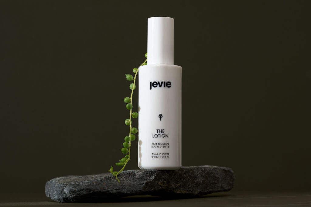 The Lotion in a white bottle on top of a grey/black stone with a green pea branch leaning on the product, going down onto the stone. On a green background.